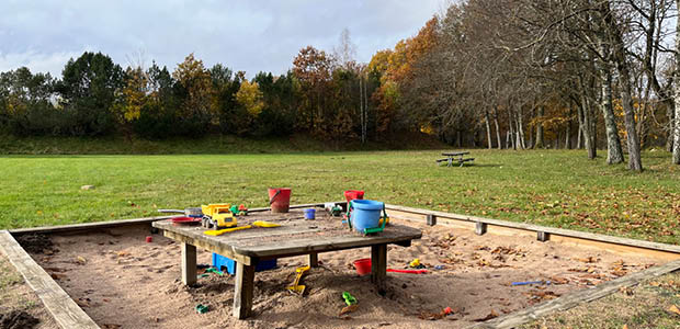 Stor sandlåda med färgglada leksaker och i bakgrunden en öppen gräsyta med höstfärgade lövträd i horisonten.